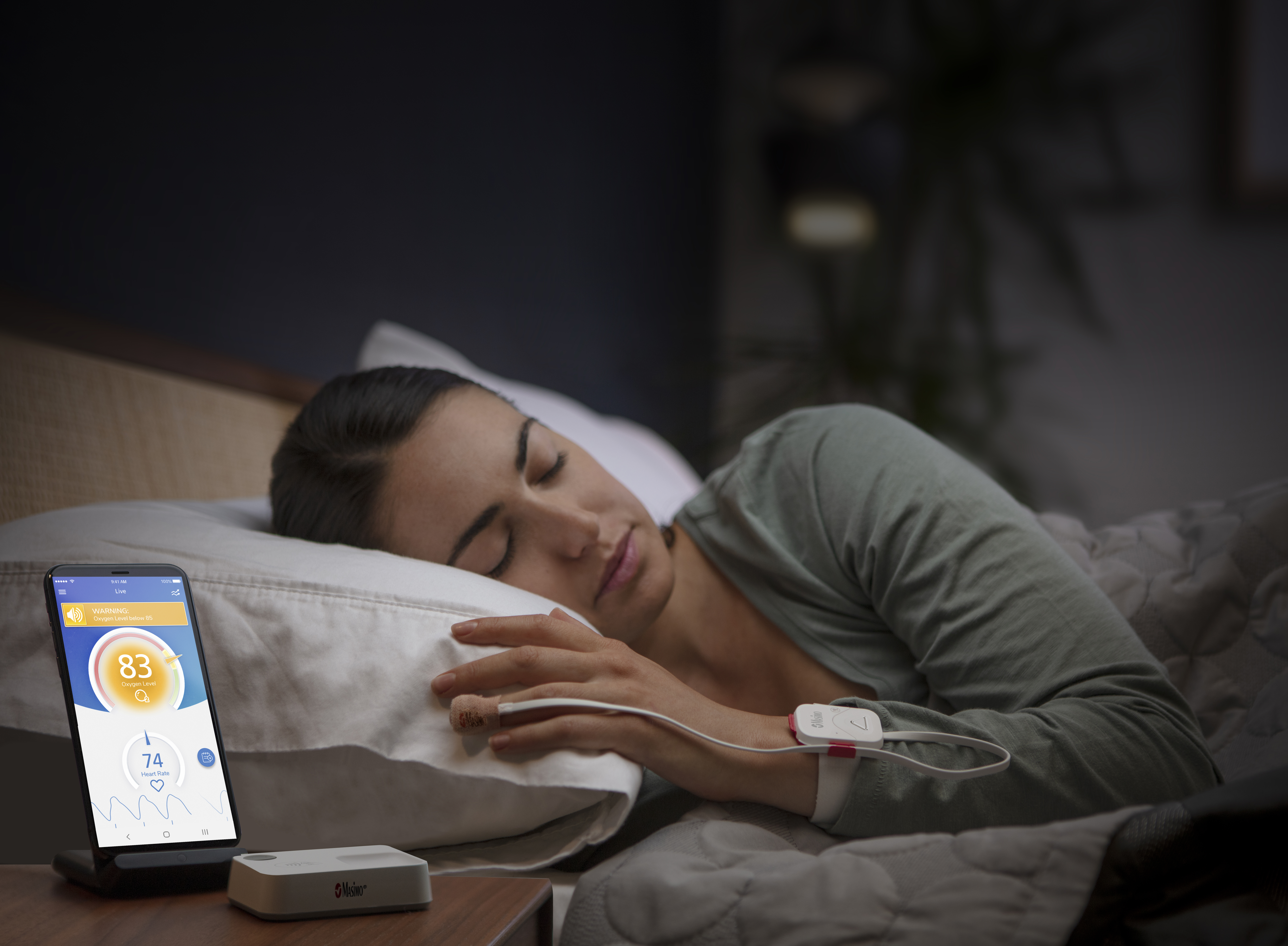 Eine Frau im Bett mit angelegtem Safetynet Alert, daneben ihr Mobiltelefon, auf dem der Bildschirm der Safetynet App anzeigt wird